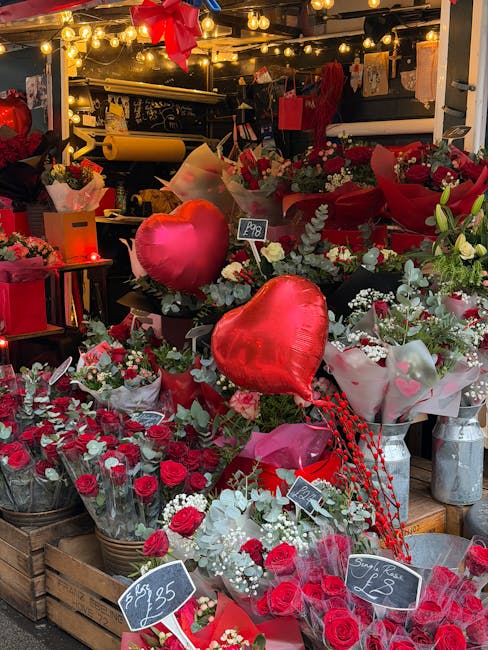 A vibrant display of fresh flowers including red roses and white blossoms arranged in baskets and wrapping paper, accented with red heart-shaped balloons and greenery. The floral arrangements are set 