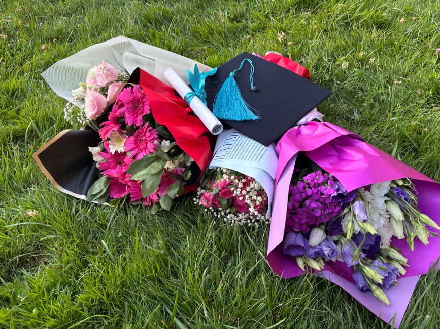 A collection of vibrant floral bouquets lies on a grassy surface, featuring pink, purple, and white flowers including roses, daisies, and lisianthus. The flowers are wrapped in colorful paper and tiss