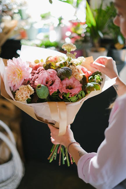 A person holding a freshly arranged floral bouquet featuring large pink peonies, soft white carnations, and delicate purple statice, accented with glossy green leaves. The bouquet is wrapped in elegan