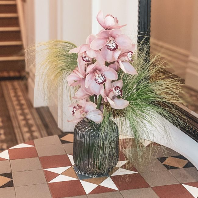 A floral arrangement featuring a tall bouquet of pink orchids with large, delicate petals and intricate white and maroon centers, placed in a dark, textured glass vase. The bouquet is accented with wi