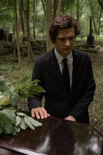 A young man in a black suit and white shirt is standing outdoors in a wooded area with tall trees and greenery in the background, looking down thoughtfully at a polished black grand piano with a bouqu