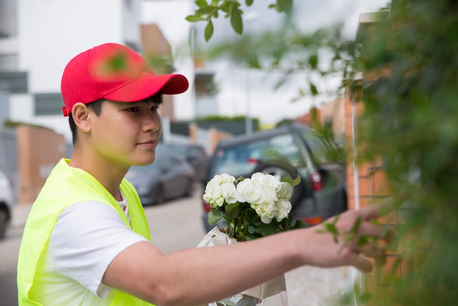 A young male florist wearing a red cap and bright yellow high-visibility vest is placing a bouquet of fresh white hydrangeas wrapped in white paper onto a doorstep. The arrangement showcases large, ro