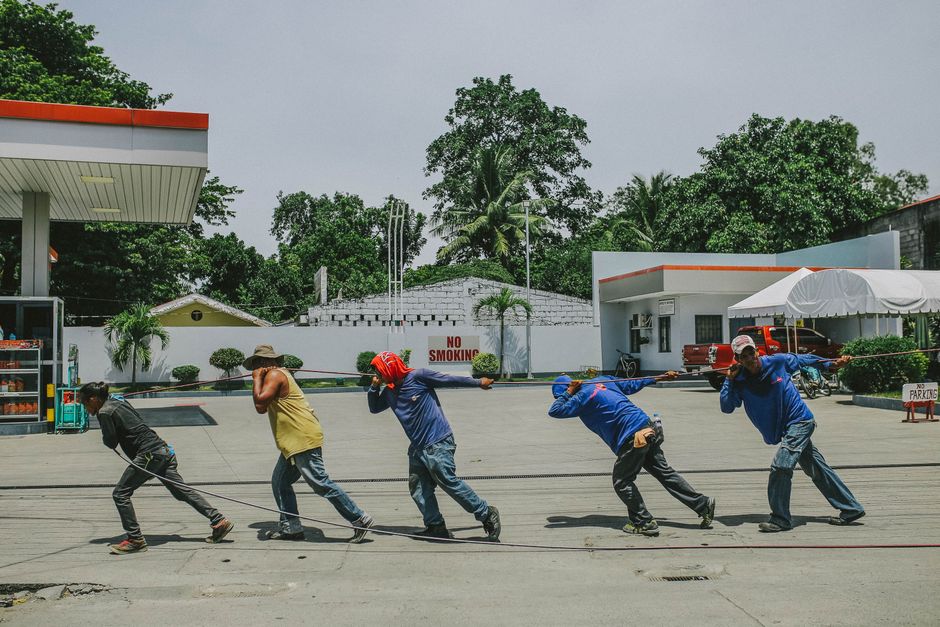 This image depicts five individuals working together to pull a long rope in an outdoor, urban setting. The group consists of diverse ages and genders, including children and adults, dressed in casual 
