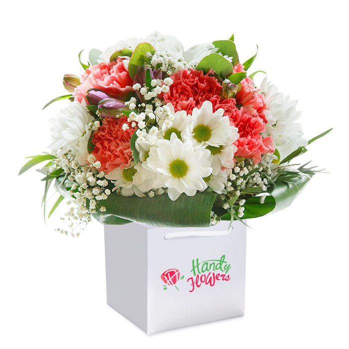 Radiating freshness and charm, this beautifully designed bouquet from Honor Oak is presented standing tall in a crisp, square white gift bag-ready for easy delivery or gifting throughout the local neighborhood. The composition features a rich blend of coral and deep red carnations, whose frilled, ruffled petals create a plush centerpiece reminiscent of summer blooms spilling over garden fences along Honor Oak Road. Cheerful white daisies with bright lime-green centers add a spirited contrast, conjuring images of wildflowers on the slopes of One Tree Hill. Fine sprays of airy white baby's breath weave delicately between the main flowers, introducing lightness and subtle movement, while a few unopened buds edged in purple and pink provide intrigue near the bouquet's left side. Lush greenery and broad, glossy leaves encircle the arrangement, forming a perfect natural frame that captures South London's love of fresh, living foliage. The expertly rounded shape reflects the skilled eye of local florists who take pride in every detail-whether crafting for an anniversary, birthday, or thoughtful pick-me-up. Against a soft white background, the bouquet's vibrant colours and artistry are emphasized, making this image a vivid illustration of the quality and care delivered to Honor Oak's homes, cafes, and creative spaces.