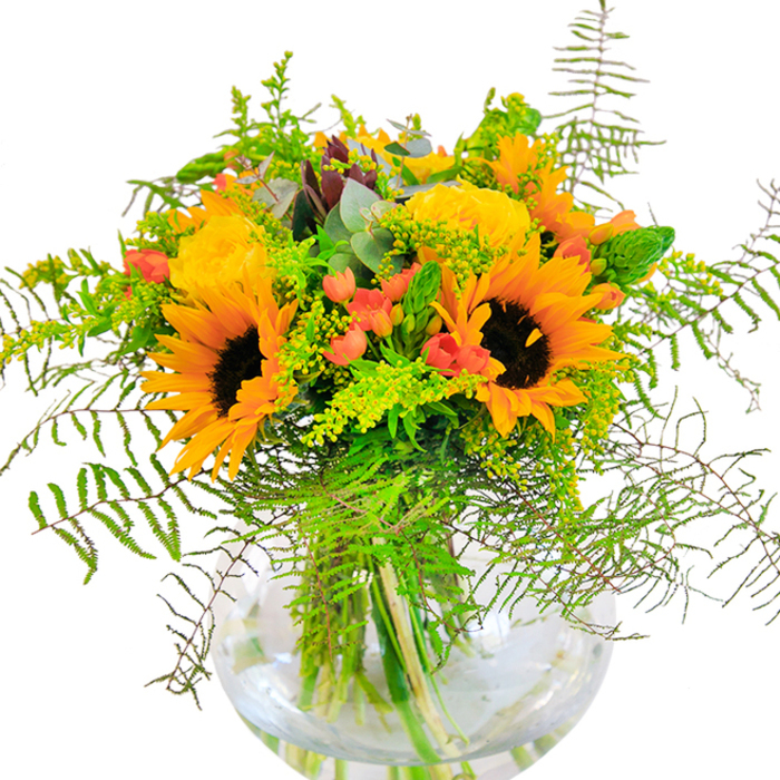 At first sight, this arrangement is a captivating burst of sunshine, reminiscent of vibrant afternoons spent beneath the sprawling canopy of One Tree Hill. Majestic sunflowers with brilliant yellow-orange petals and rich, chocolate centers form the visual heart, bringing the energy of summer's zenith to the bouquet. Between their impressive heads, garden roses in soft shades of golden yellow spiral open, their velvety textures recalling the cultivated blooms from local Honor Oak gardens. Glossy, deep red Hypericum berries glisten among the foliage, their round, lively shapes and fiery hue providing perfect punctuation to the composition-like gems sparkling in the light that filters through Stondon Park. Fern fronds, graceful and feathery, cascade outward, while light green filler and slender flower spikes add gentle movement and natural fullness. Subtle notes of blue-green from eucalyptus leaves peek between the florals, conjuring a breath of coolness akin to that found after summer rainfall. The whole ensemble is artfully displayed in a clear, globe-shaped vase, the clean lines and water reflecting every stem's fresh vigour. This bouquet's construction-abundant and harmonious-speaks to the pride and expertise of Honor Oak's florists, making it perfect for celebrating notable milestones, surprising a loved one, or elevating an everyday moment in the comfort of an SE23 home.